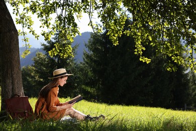 Photo of Young woman reading book under tree on meadow in mountains