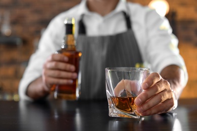 Bartender with glass and bottle of whiskey at counter in bar, closeup. Space for text