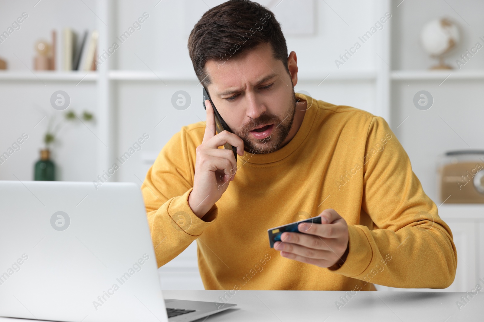 Photo of Upset man with credit card talking on smartphone near laptop at home. Be careful - fraud