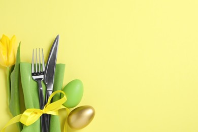 Photo of Cutlery set, painted eggs and beautiful flower on yellow background, flat lay with space for text. Easter celebration