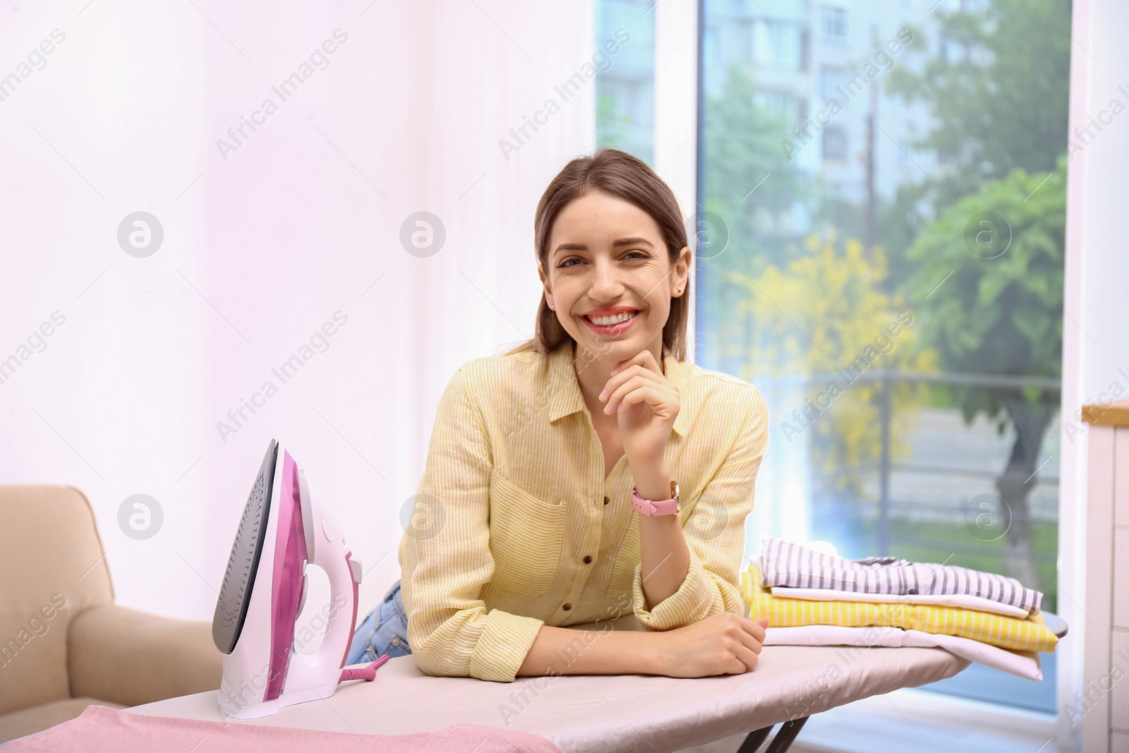 Photo of Young pretty woman with iron and clean laundry near board indoors