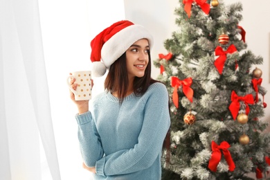 Beautiful young woman in Santa hat with cup of drink near Christmas tree at home