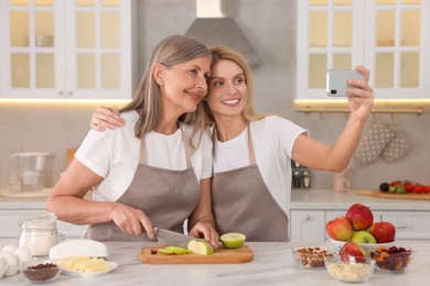 Photo of Happy daughter taking selfie with her mature mother while cooking together in kitchen