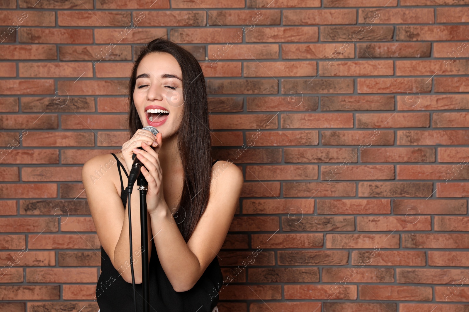 Photo of Young stylish woman singing in microphone near brick wall. Space for text