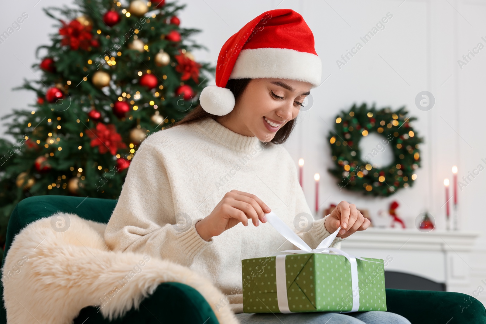 Photo of Happy young woman in Santa hat opening gift box in room decorated for Christmas