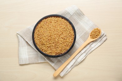 Bowl and spoon with uncooked bulgur on wooden table, flat lay