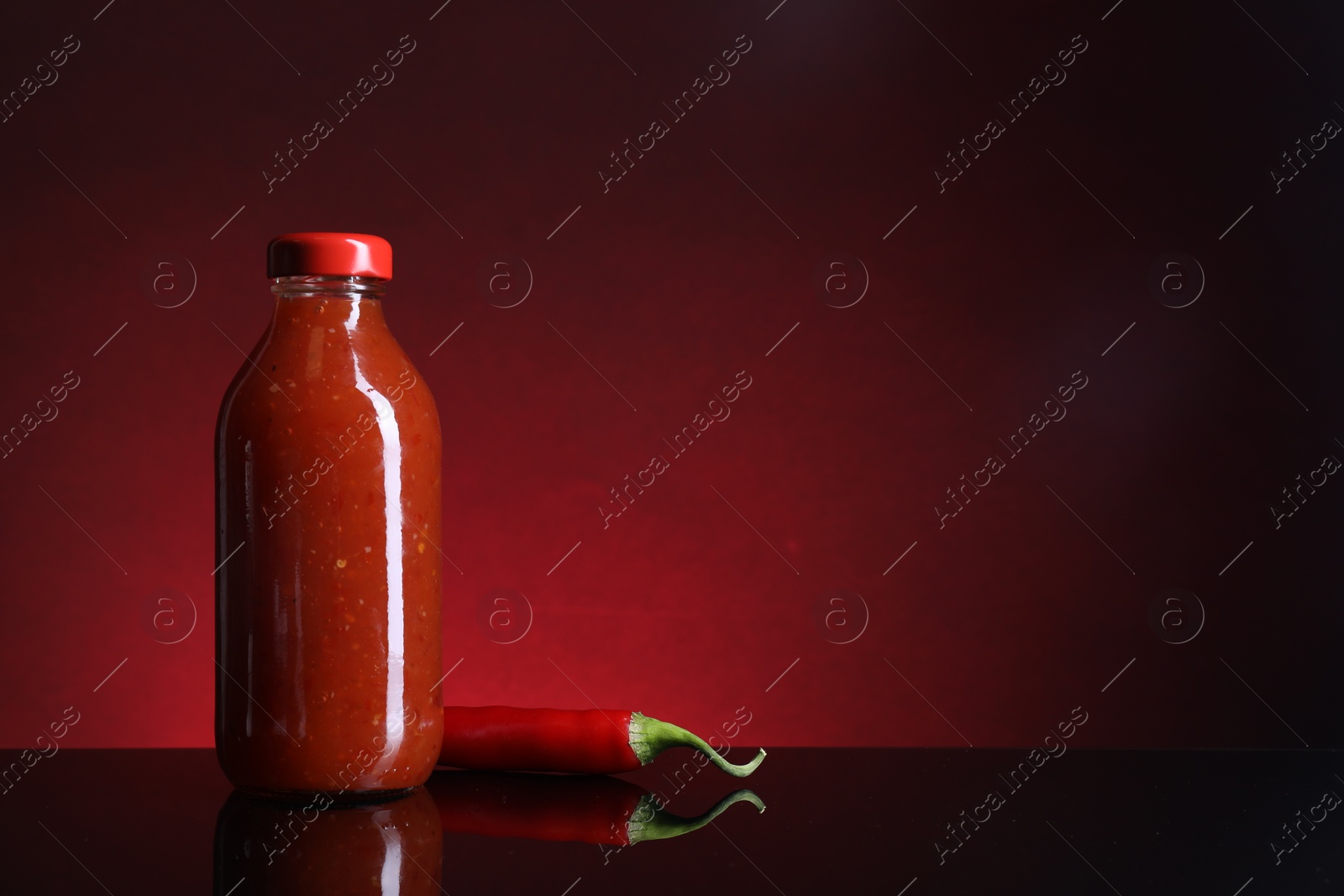 Photo of Spicy chili sauce in bottle and pepper against dark background, space for text