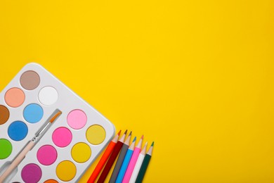 Photo of Watercolor palette with brush and colorful pencils on yellow background, flat lay. Space for text