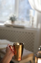 Woman with golden cup of hot drink in room decorated for Christmas, closeup