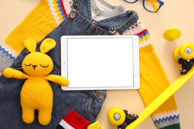 Photo of Modern tablet, clothes, skateboard and toy bunny on beige background, flat lay. Space for text