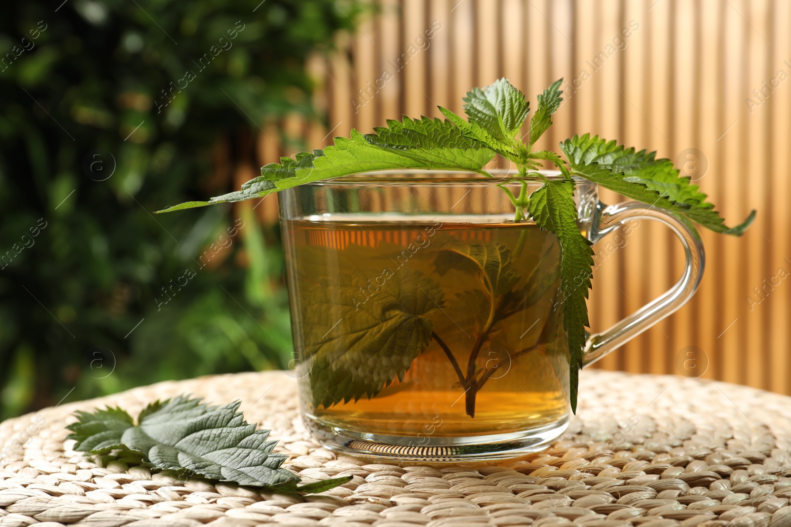 Photo of Glass cup of aromatic nettle tea and green leaves on wicker mat, closeup