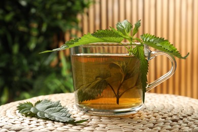 Photo of Glass cup of aromatic nettle tea and green leaves on wicker mat, closeup