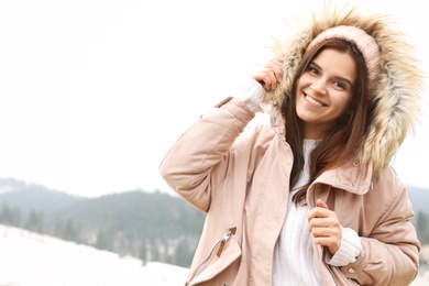 Young woman in warm clothes near snowy hill, space for text. Winter vacation