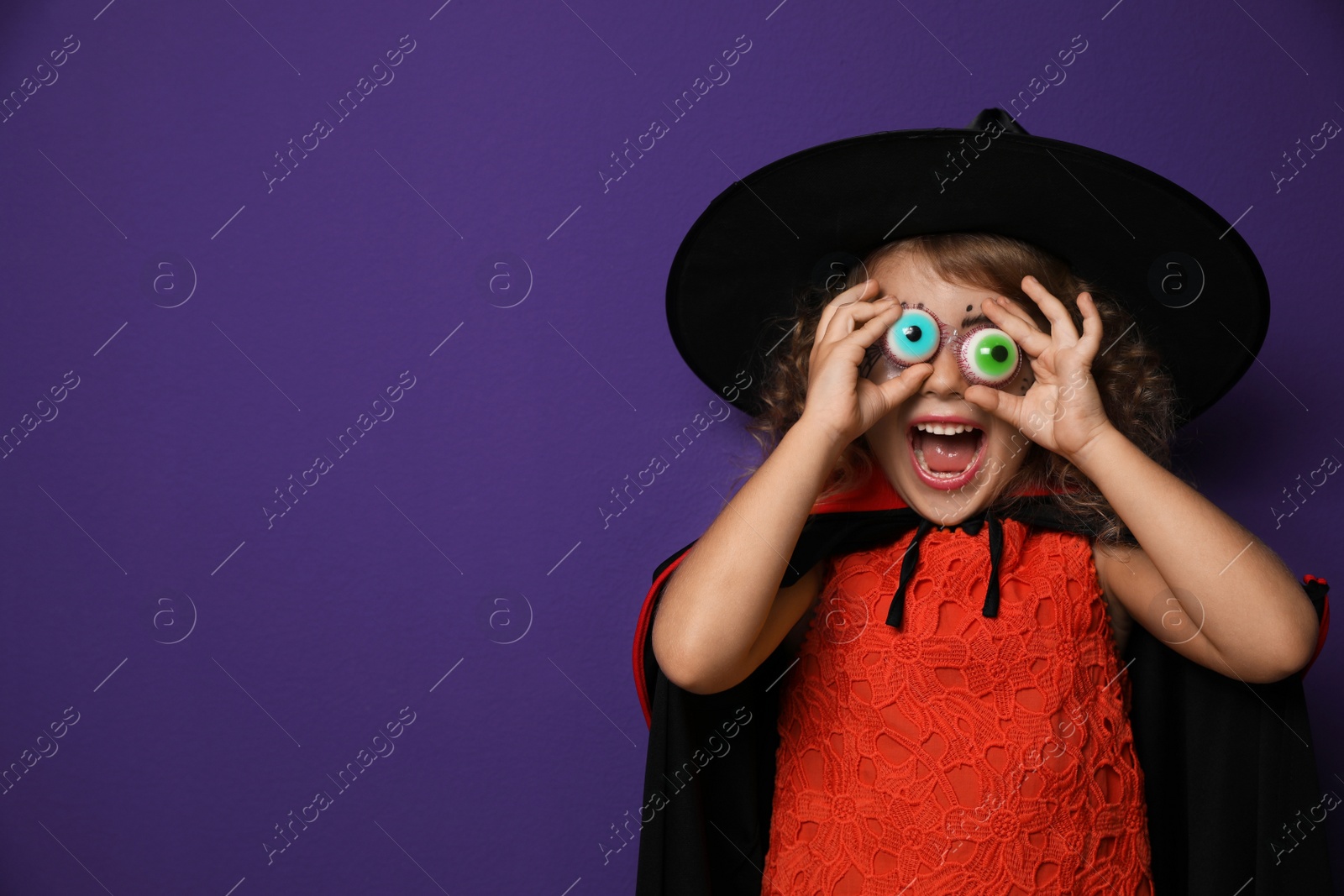 Photo of Cute little girl with toy eyes wearing Halloween costume on purple background. Space for text