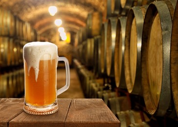 Glass of tasty beer on wooden table in cellar with large barrels, space for text