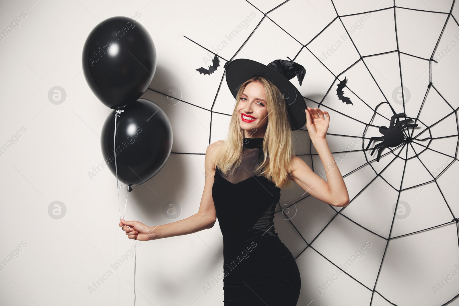 Photo of Woman in witch hat with balloons posing near white wall decorated for Halloween