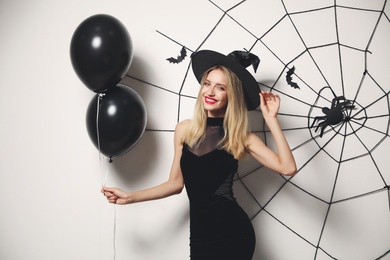 Woman in witch hat with balloons posing near white wall decorated for Halloween