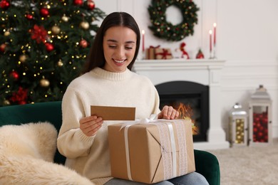 Happy young woman with Christmas gift reading greeting card at home