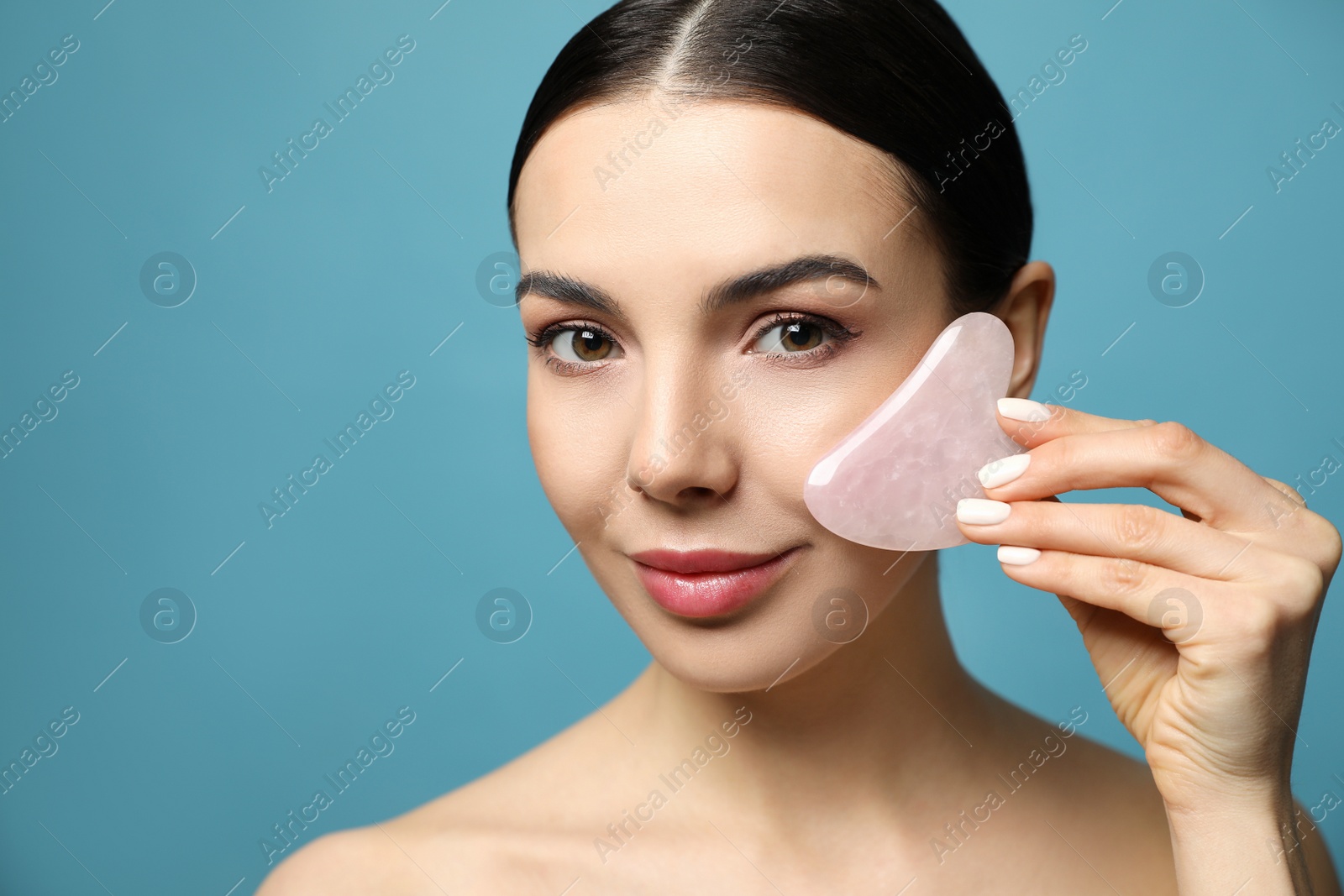 Photo of Beautiful young woman doing facial massage with gua sha tool on blue background, closeup