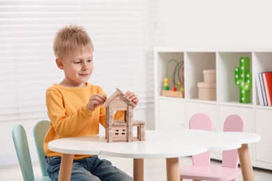 Cute little boy playing with wooden house at white table indoors, space for text. Child's toy
