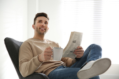 Photo of Young man with cup of drink and newspaper relaxing in armchair near window at home