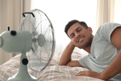 Photo of Man enjoying air flow from fan on bed in room. Summer heat