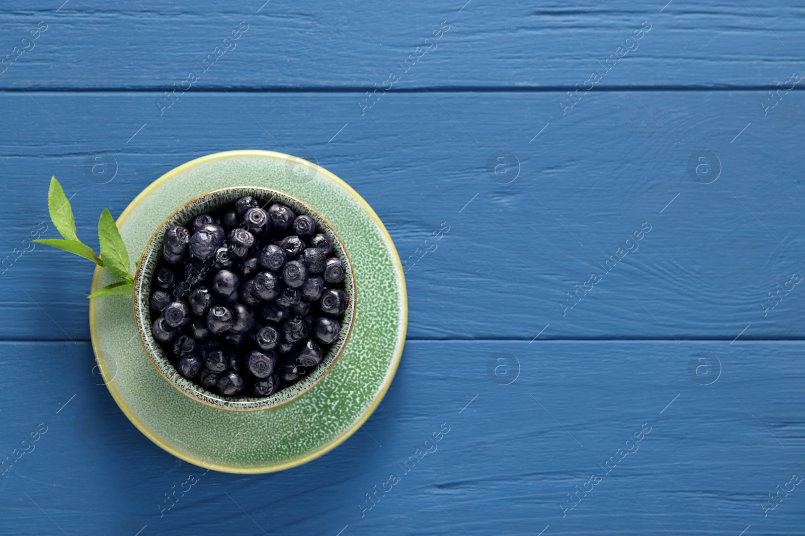 Photo of Ripe bilberries in bowl and sprig on blue wooden table, top view. Space for text