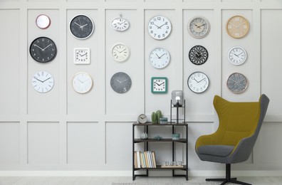 Armchair, console table and many different clocks hanging on white wall in room