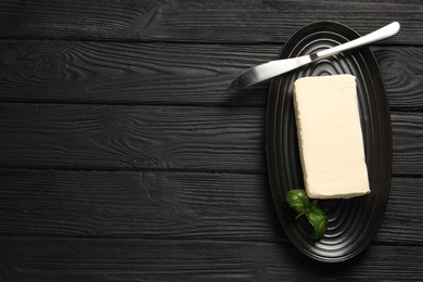 Photo of Block of tasty butter with basil and knife on black wooden table, top view. Space for text