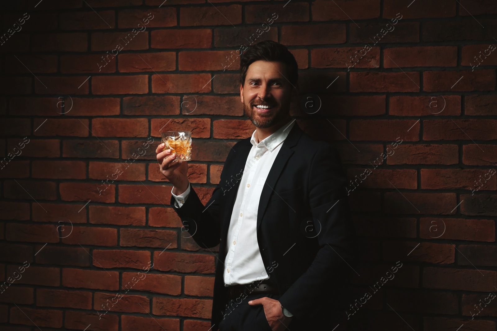 Photo of Man in formal suit holding glass of whiskey near red brick wall