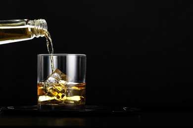 Pouring whiskey from bottle into glass with ice cubes at table against black background, space for text