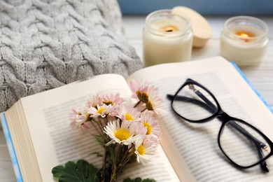 Photo of Open book with chamomile flowers as bookmark, scented candle and glasses on table, closeup