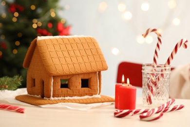 Photo of Tasty gingerbread house and burning candles on light table indoors