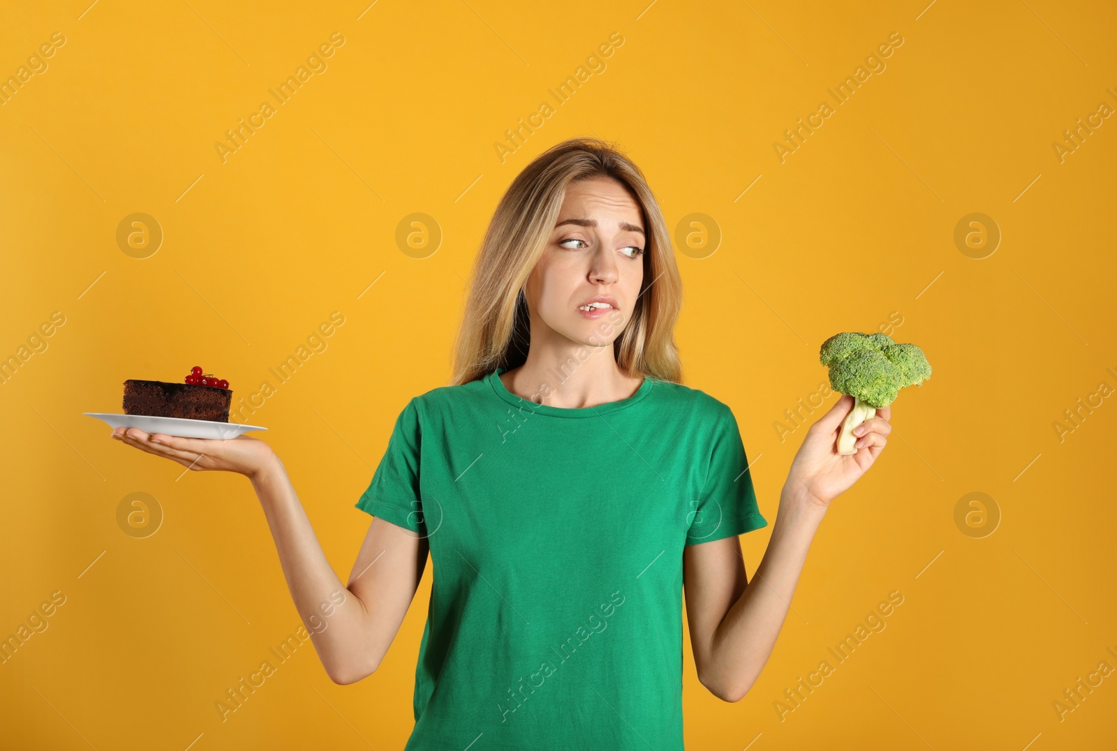 Photo of Woman choosing between cake and healthy broccoli on yellow background