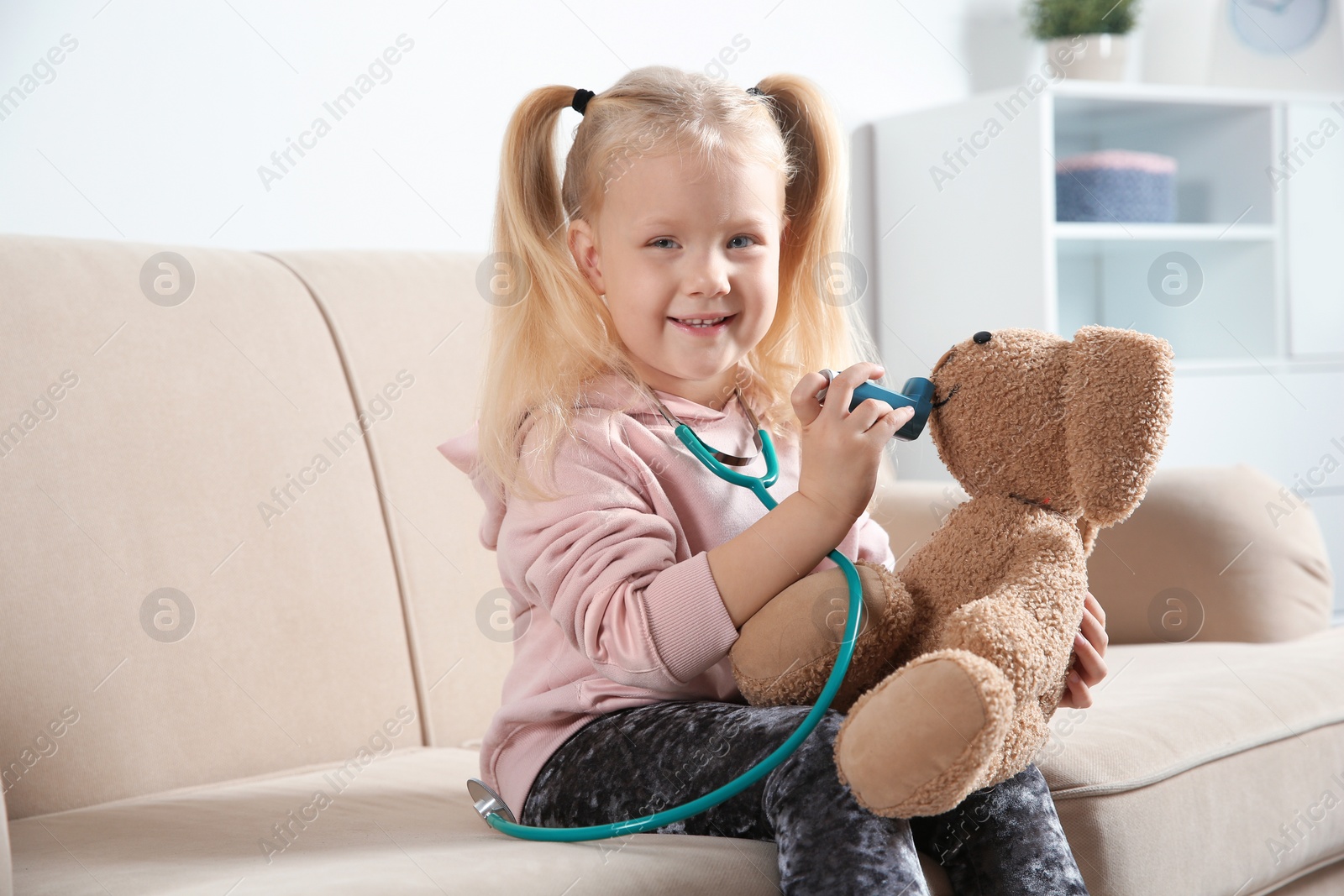 Photo of Cute child imagining herself as doctor while playing with toy bunny on couch at home