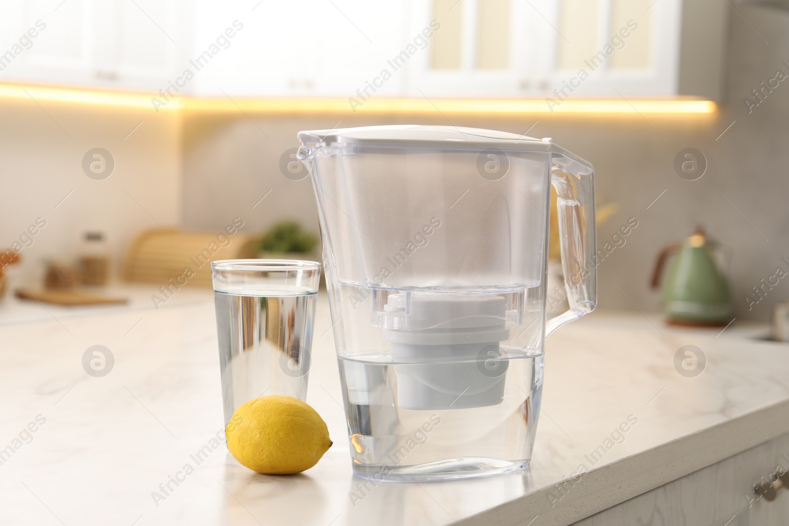 Photo of Water filter jug, glass and lemon on white marble table in kitchen