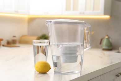 Photo of Water filter jug, glass and lemon on white marble table in kitchen