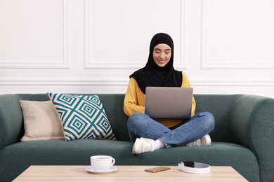 Photo of Muslim woman in hijab using laptop on sofa indoors. Space for text