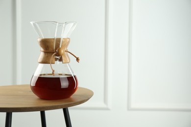 Glass chemex coffeemaker with coffee on wooden table against white wall, space for text