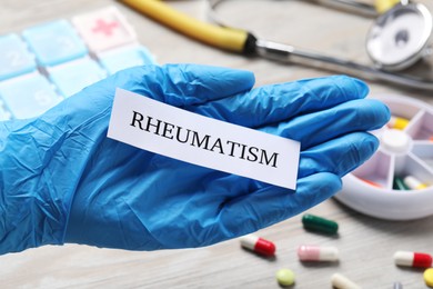 Doctor in glove holding sheet of paper with word Rheumatism above table with pills, closeup