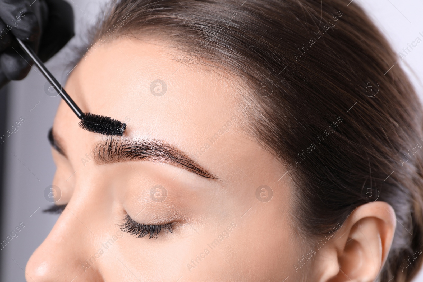 Photo of Beautician brushing woman's eyebrows after tinting on light background, closeup