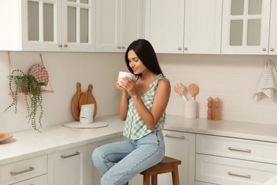Beautiful young woman with cup of tea sitting on stool in kitchen