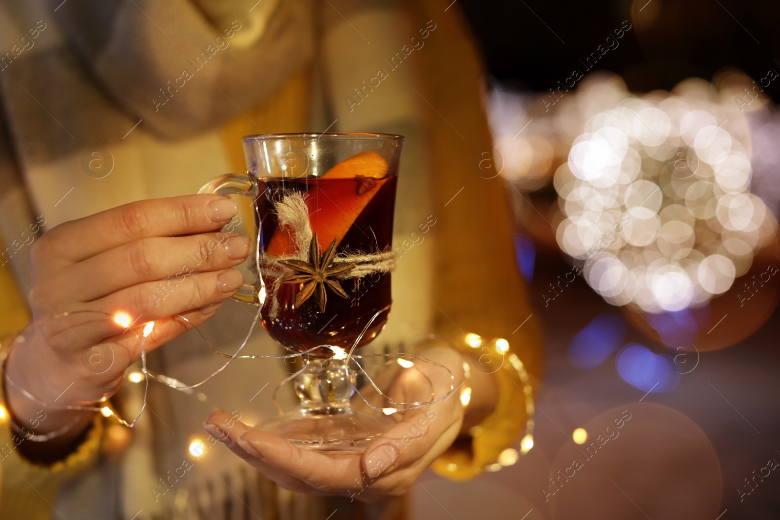 Photo of Woman with glass cup of mulled wine and garland at winter fair, closeup. Space for text