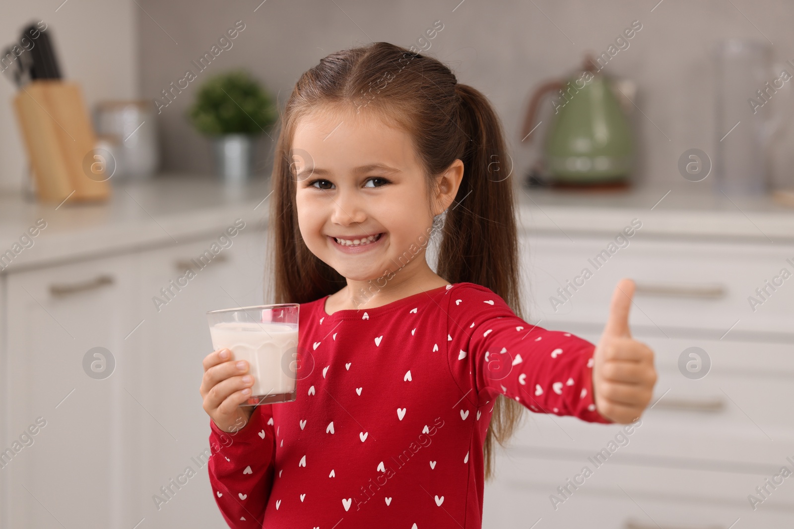 Photo of Cute girl with glass of fresh milk showing thumb up in kitchen