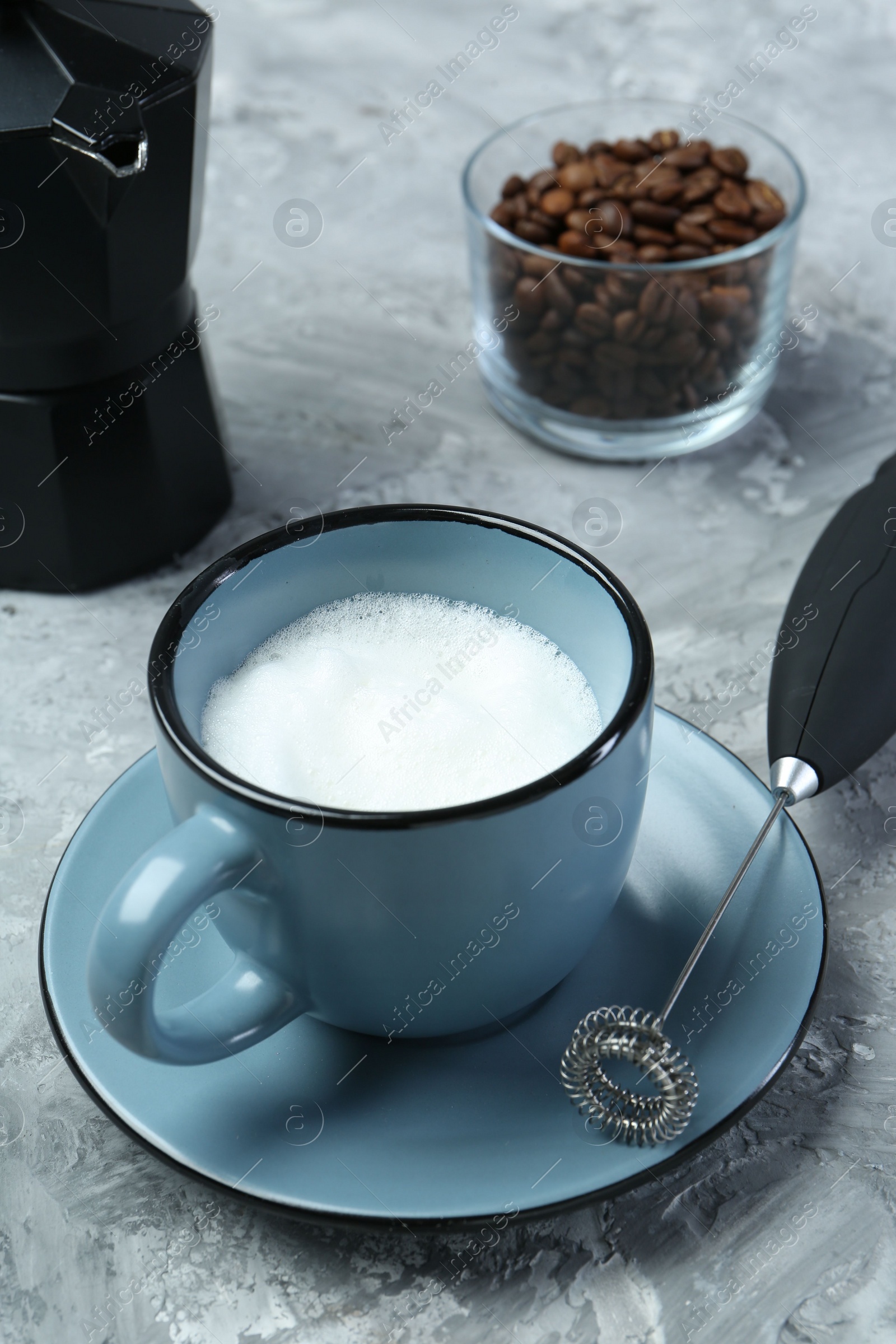 Photo of Mini mixer (milk frother), whipped milk in cup and coffee beans on grey textured table