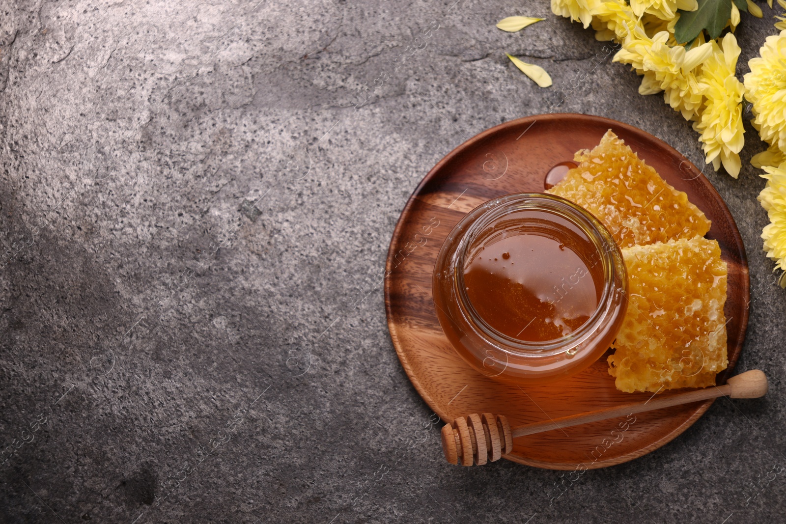 Photo of Sweet golden honey in jar, dipper, pieces of honeycomb and chrysanthemum flowers on grey textured table, flat lay. Space for text