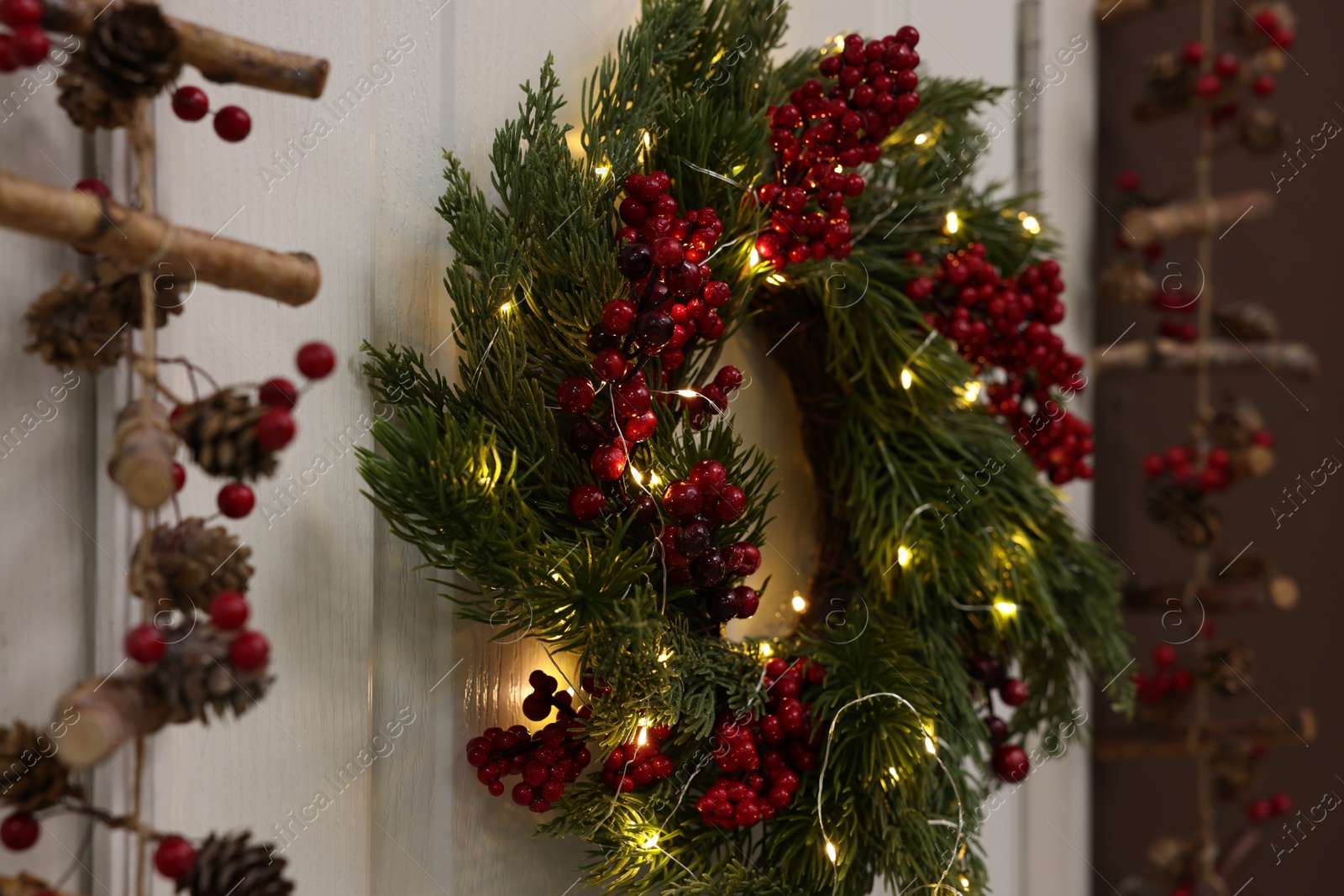 Photo of Beautiful Christmas wreath with red berries and fairy lights hanging on white door, closeup. Space for text