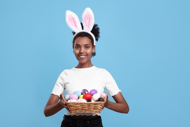Happy African American woman in bunny ears headband holding wicker tray with Easter eggs on light blue background