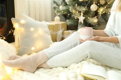 Photo of Woman with cup of coffee and book sitting on soft plaid indoors, closeup. Christmas celebration