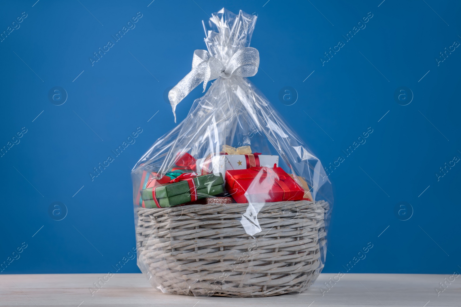 Photo of Wicker basket full of gift boxes on white wooden table against blue background